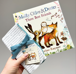 Hand holding out story print dribble cloths tied in turquoise ribbon and matching book, Catherine Rayner's 'Molly, Olive and Dexter: Three Best Friends.'