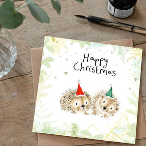 Christmas card with hedgehogs wearing hats on a wooden surface with stationery items.