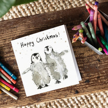 Load image into Gallery viewer, A Christmas card featuring two baby penguins on a wooden board with colour pencils around it