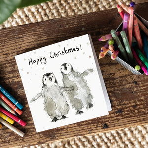 A Christmas card featuring two baby penguins on a wooden board with colour pencils around it