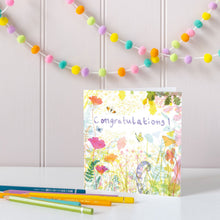 Load image into Gallery viewer, Greeting card with floral design and 'Congratulations!' text, placed on a white surface with colorful pencils and a string of multicolored pom-poms in the background.