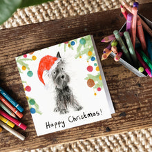 Load image into Gallery viewer, A square business card lying on a wooden board with colour pencils featuring a Scottish terrier in a red Santa hat surrounded with Christmas tree branches with baubles on them.