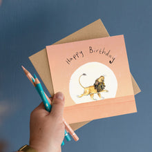 Load image into Gallery viewer, Hand holding Happy Birthday Card featuring an illustration of a Lion walking across a pink sunset, as well as the pencils used to draw the greetings card.