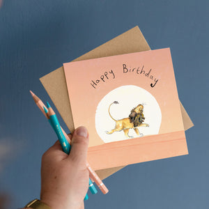 Hand holding Happy Birthday Card featuring an illustration of a Lion walking across a pink sunset, as well as the pencils used to draw the greetings card.