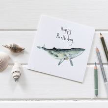 Load image into Gallery viewer, White birthday card featuring watercolour whale illustration surrounded by shells and pencil crayons on wooden decking. 