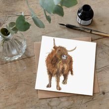Load image into Gallery viewer, Highland cow greetings card lying on a wooden desk beside an ink bottle and dip pen. 