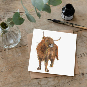 Highland cow greetings card lying on a wooden desk beside an ink bottle and dip pen. 