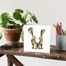 Load image into Gallery viewer, Greetings hard featuring an expressive watercolour illustration of two hares back to back on a bench beside a box of colouring pencils.