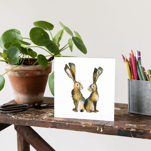 Greetings hard featuring an expressive watercolour illustration of two hares back to back on a bench beside a box of colouring pencils.