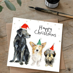 Illustrated christmas card featuring 3 dogs wearing santa hats lying on a wooden desk with a dip pen and matching brown envelope. 